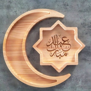 May include: Wooden serving tray in the shape of a crescent moon and an eight-pointed star. The star-shaped tray has Arabic calligraphy etched into the wood. The wood has a light, natural finish.