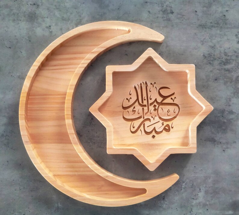 May include: Wooden serving tray in the shape of a crescent moon and an eight-pointed star. The star-shaped tray has Arabic calligraphy etched into the wood. The wood has a light, natural finish.