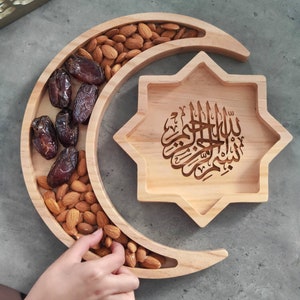 May include: Wooden serving tray in a crescent moon and star shape. The crescent moon section holds almonds and dates. The star-shaped section features Arabic calligraphy. The tray is made of light-colored wood.