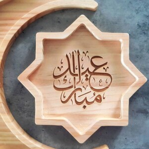 May include: A wooden star-shaped tray with carved Arabic calligraphy, resting on a crescent-shaped wooden base. The tray is light brown, and the calligraphy is in a darker shade. The text reads "Eid Mubarak".