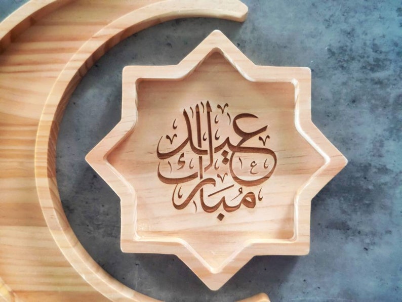 May include: A wooden star-shaped tray with carved Arabic calligraphy, resting on a crescent-shaped wooden base. The tray is light brown, and the calligraphy is in a darker shade. The text reads "Eid Mubarak".