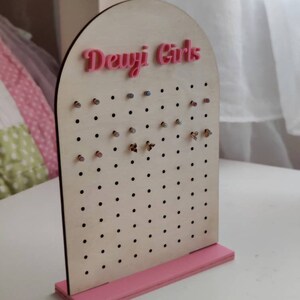 May include: A wooden earring display stand with a pink base and the text "Dewji Girls" in pink. The display has multiple holes for earrings and is made of light-coloured wood.