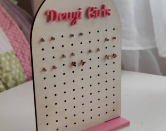 May include: A wooden earring display stand with a pink base and the text "Dewji Girls" in pink. The display has multiple holes for earrings and is made of light-coloured wood.