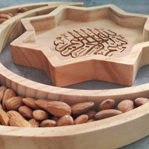 May include: A wooden serving tray with a star-shaped center and a crescent-shaped outer ring. The star-shaped section features engraved Arabic calligraphy. The crescent and star sections are filled with almonds.