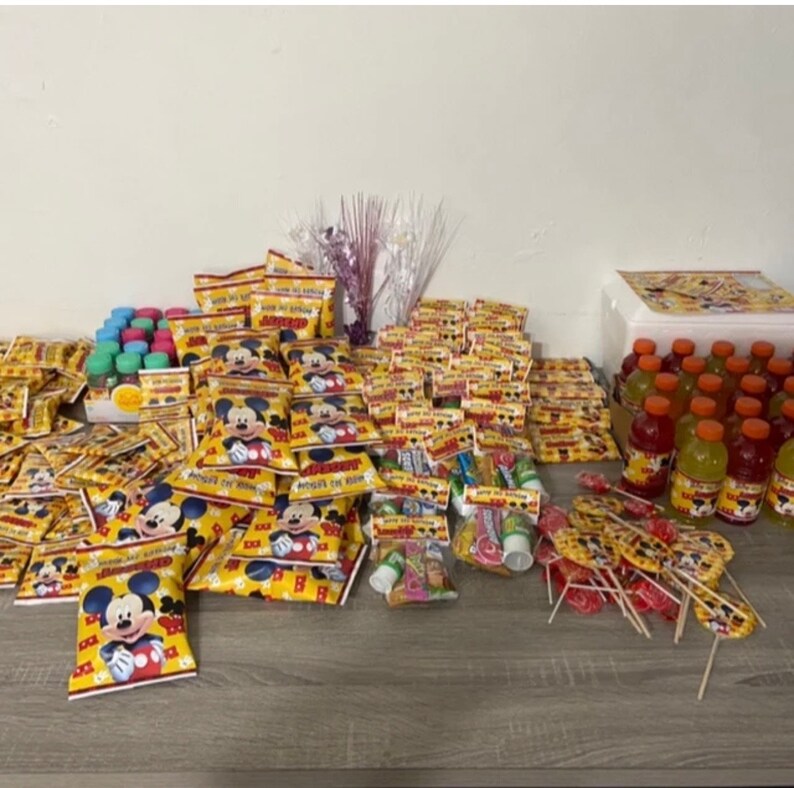 May include: A variety of party favors for a Mickey Mouse themed birthday party. The favors include small bags of chips, lollipops, juice boxes, and bubble wands. The bags of chips have a Mickey Mouse design and the lollipops are yellow and red with a Mickey Mouse design. The juice boxes are red and orange with a Mickey Mouse design. The bubble wands are silver and have a Mickey Mouse design.