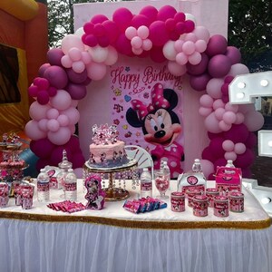 May include: A pink and white balloon arch with a Minnie Mouse backdrop and a cake decorated with a crown. There are also small containers of sweets and a light-up number 3.
