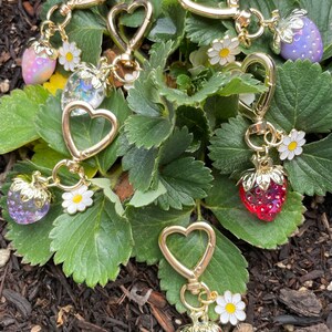 Puede incluir: Cinco llaveros de coraz&oacute;n dorado con dijes de fresa y detalles de flores blancas. Los dijes son de varios colores, incluyendo rojo, rosa y morado.