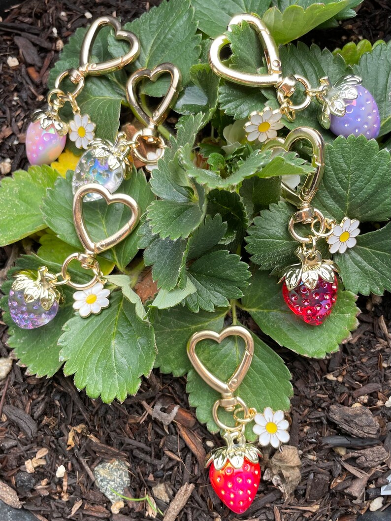 Puede incluir: Cinco llaveros de coraz&oacute;n dorado con dijes de fresa y detalles de flores blancas. Los dijes son de varios colores, incluyendo rojo, rosa y morado.