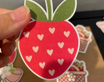 May include: A red strawberry sticker with white heart-shaped accents. The sticker has green glittery leaves and a white stem. Other stickers with strawberries and baskets are in the background.