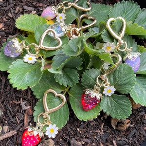 Puede incluir: Un conjunto de cinco llaveros de tono dorado con dijes de fresa. Cada dije es de un color diferente, incluyendo rojo, rosa, morado y transparente. Los llaveros est&aacute;n unidos a anillos de coraz&oacute;n de tono dorado.