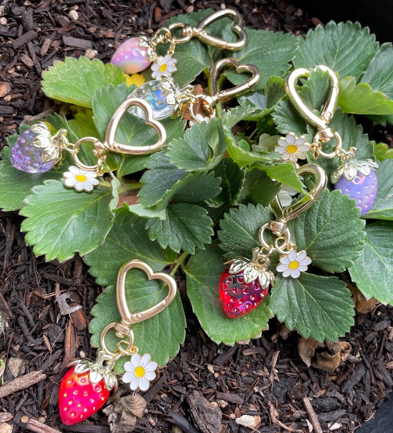 Puede incluir: Un conjunto de cinco llaveros de tono dorado con dijes de fresa. Cada dije es de un color diferente, incluyendo rojo, rosa, morado y transparente. Los llaveros est&aacute;n unidos a anillos de coraz&oacute;n de tono dorado.