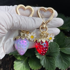 Puede incluir: Dos llaveros de coraz&oacute;n de oro con un dije de fresa. Una fresa es roja y la otra es p&uacute;rpura iridiscente. Cada fresa tiene una hoja de oro y un dije de margarita blanca.