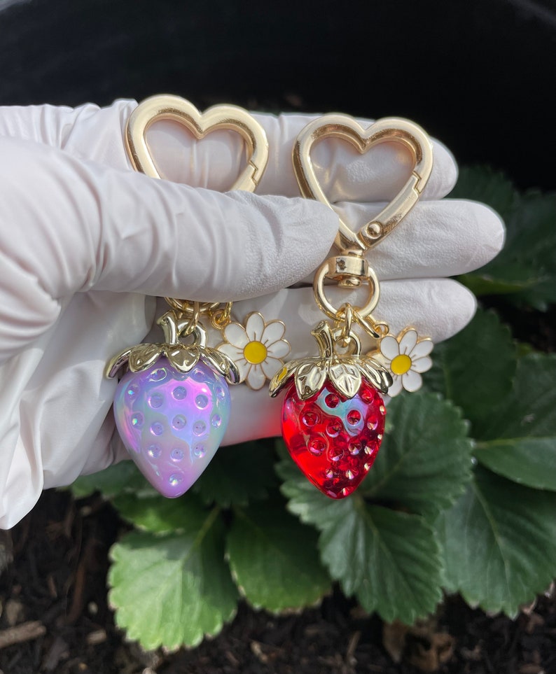 Puede incluir: Dos llaveros de coraz&oacute;n de oro con un dije de fresa. Una fresa es roja y la otra es p&uacute;rpura iridiscente. Cada fresa tiene una hoja de oro y un dije de margarita blanca.