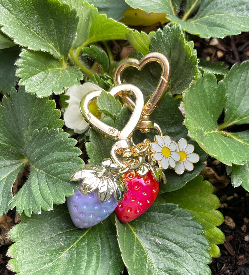 Puede incluir: Un llavero de encanto dorado con dos encantos de fresa, uno rojo y otro morado iridiscente, y un encanto de margarita blanca.