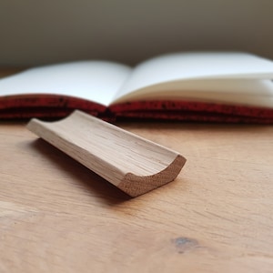 May include: A wooden pen holder with a curved shape, made of light brown wood. The pen holder is sitting on a wooden surface.