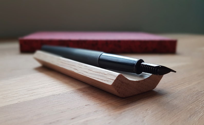 May include: A black fountain pen with a metal nib resting in a wooden pen holder. The holder is made of light wood with a natural grain pattern.