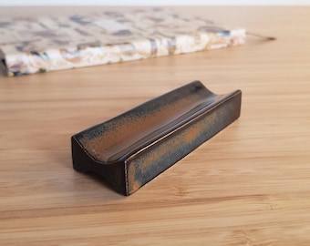 May include: A rectangular, dark brown ceramic soap dish with a curved interior. The dish has a textured, aged appearance with hints of rust and sits on a light brown wooden surface. A patterned notebook is in the background.