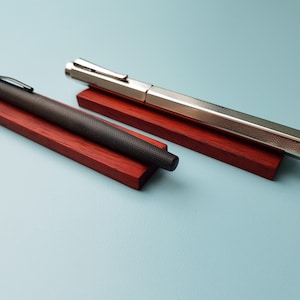 May include: Two pens on wooden stands. The pen on the left is black with a textured finish. The pen on the right is silver with a textured finish. Both pens are on red wooden stands.