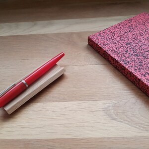 May include: A red fountain pen with a silver trim is resting on a wooden pen holder. The pen is on a wooden surface next to a red notebook with black speckles.