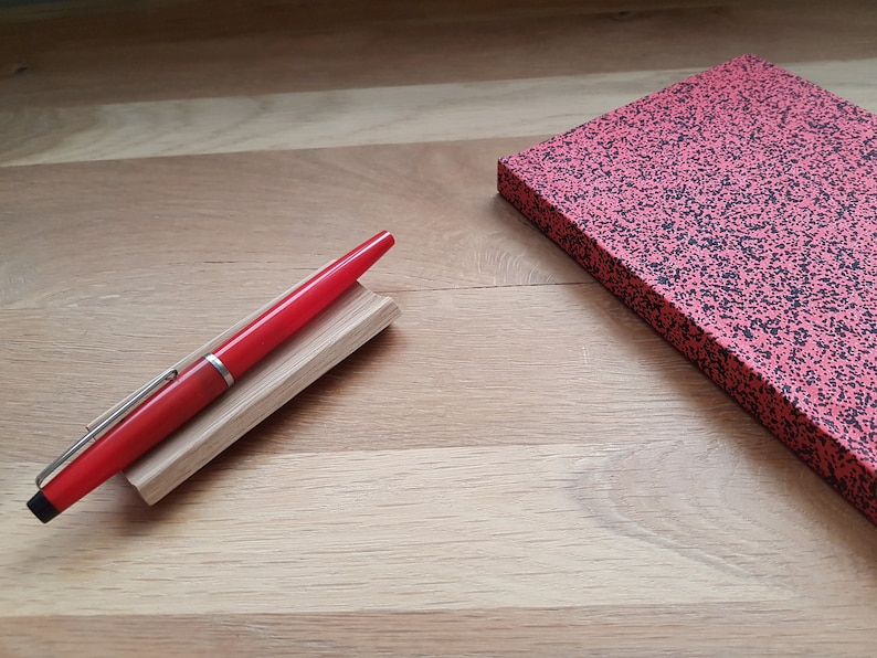 May include: A red fountain pen with a silver trim is resting on a wooden pen holder. The pen is on a wooden surface next to a red notebook with black speckles.