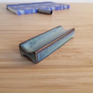 Op de afbeelding: Een rechthoekige keramische asbak met een turquoise glazuur en bronzen randen. De asbak heeft een gebogen ontwerp en ligt op een houten oppervlak. Een blauw notitieboek en een metalen pen zijn op de achtergrond.