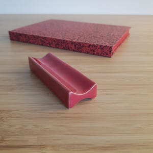 Può includere: Un poggia cucchiaio in ceramica rossa con un design curvo è posto di fronte a un sottobicchiere rettangolare rosso maculato. Gli oggetti sono su una superficie di legno marrone chiaro. Il poggia cucchiaio è di colore rosso lucido.