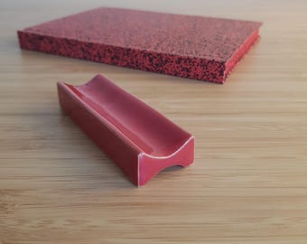 May include: A red ceramic spoon rest with a curved design sits in front of a rectangular red speckled coaster. The items are on a light brown wooden surface. The spoon rest is a solid, glossy red color.