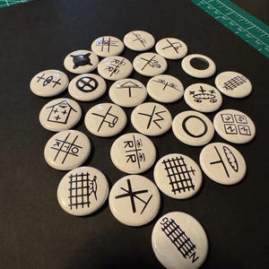 May include: A collection of white, circular buttons with various black graphic designs. The designs include geometric shapes, symbols, and abstract patterns. The buttons are arranged on a black surface, with a green cutting mat in the background.