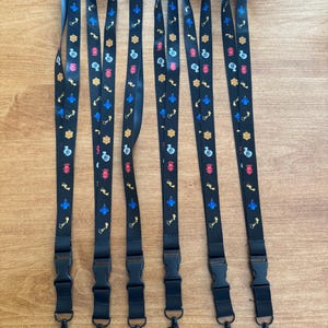May include: Six black lanyards with colorful repeating patterns of various shapes and symbols. Each lanyard has a black plastic clasp and a metal hook. The lanyards are lying on a light brown wooden surface.