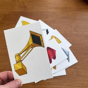 May include: A hand holds a stack of white cards with illustrations of various objects. The top card features a yellow and black gramophone illustration. Other cards show a red hat, a yellow horn, and other objects.