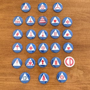 May include: A collection of thirty circular pin-back buttons featuring blue backgrounds with white triangles containing various red symbols, including letters like CD, CDC, and E, alongside icons such as a flame, ladder, cross, tree, and steering wheel.