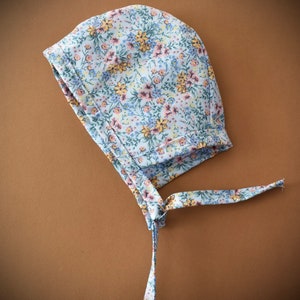May include: A white bonnet with a floral print featuring yellow, pink, and blue flowers. The bonnet has a long tie that can be tied under the chin.