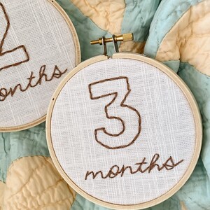 May include: Two white embroidery hoops with brown thread. The first hoop says "2 months" and the second hoop says "3 months".
