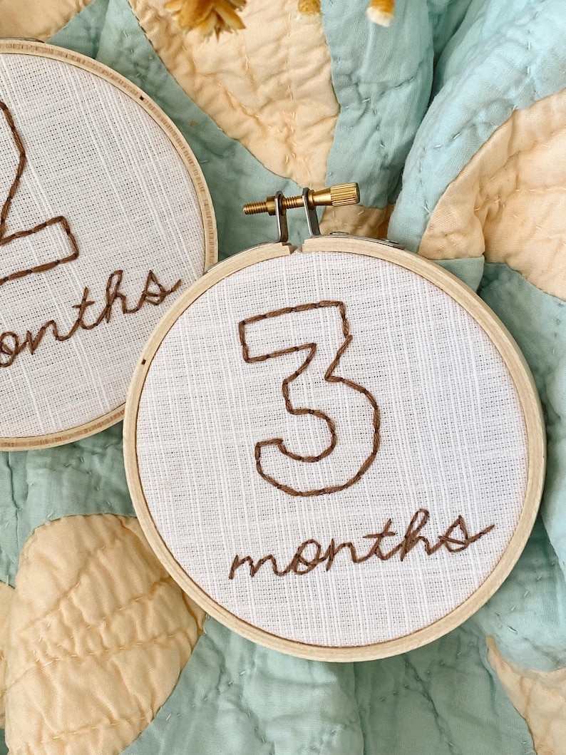 May include: Two white embroidery hoops with brown thread. The first hoop says "2 months" and the second hoop says "3 months".