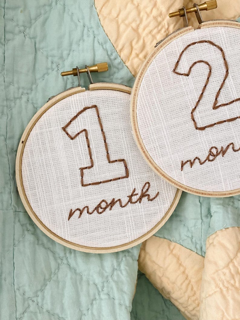 May include: Two white fabric circles with brown embroidery. The first circle has the number "1" and the text "month". The second circle has the number "2" and the text "month".