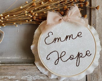 May include: A white fabric embroidery hoop with the names "Emme Hope" stitched in brown thread. A light pink bow is attached at the top. The hoop is set on a white, scalloped fabric, with dried flowers in the background.