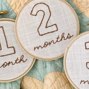 May include: Three white fabric circles with brown embroidery. The circles are numbered 1, 2, and 3 and each has the word "months" embroidered below the number.