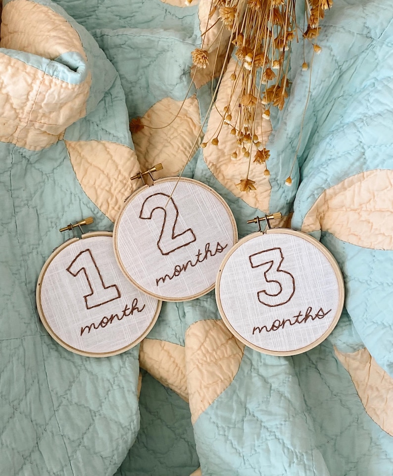 May include: Three embroidered hoop ornaments with the numbers 1, 2, and 3, each with the word "month" or "months" written below. The hoops are on a light blue and cream quilt.