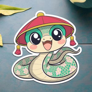 Peut inclure: Un serpent de dessin animé portant un chapeau rouge et or avec un pompon. Le serpent a des yeux verts, un nez rose et un corps à motifs vert clair et rose.