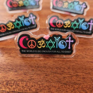 May include: A clear acrylic pin with a black background and colorful religious symbols. The text "The world is big enough for all to exist" is printed on the pin.