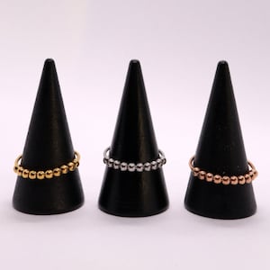May include: Three beaded rings on black cone-shaped display stands. The rings are gold, silver, and rose gold.