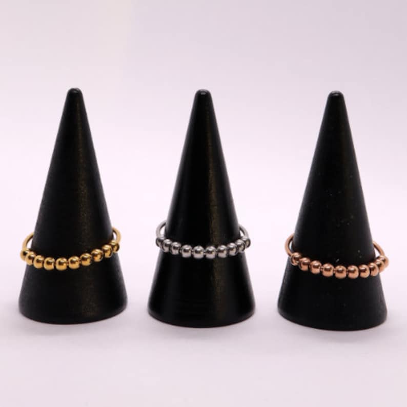 May include: Three beaded rings on black cone-shaped display stands. The rings are gold, silver, and rose gold.