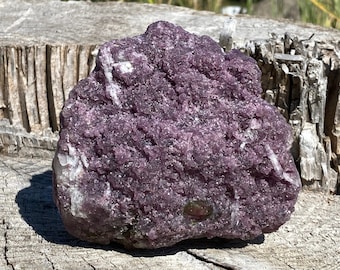 May include: A rough, uncut piece of purplish-pink lepidolite mineral, with white and clear crystalline inclusions. The stone is displayed on a weathered wooden surface, with a backdrop of green foliage and a clear blue sky.