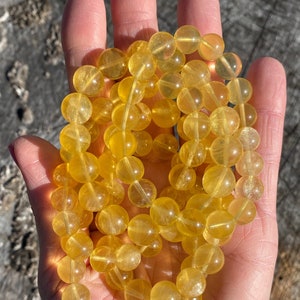 May include: A strand of golden yellow gemstone beads, each approximately 0.5 inches in diameter, held in a hand. The beads have a translucent quality, catching the light. The overall theme is jewelry and gemstones.