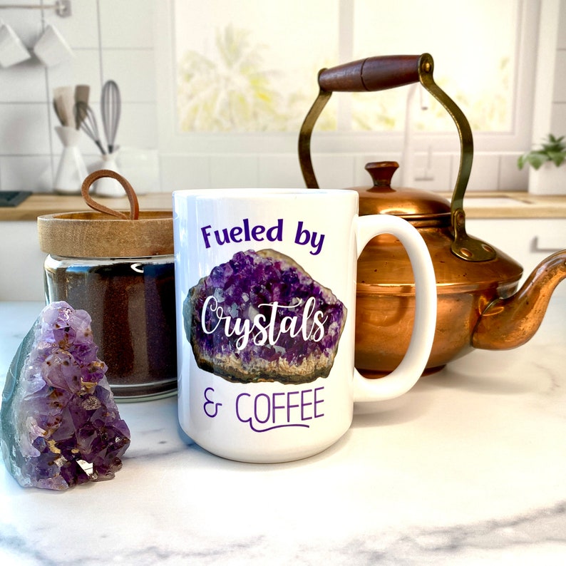 Peut inclure: Une tasse &agrave; caf&eacute; en c&eacute;ramique blanche avec un motif de cristal d'am&eacute;thyste violet et le texte "Fueled by Crystals & Coffee".