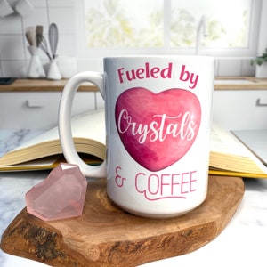 May include: White ceramic coffee mug with a pink heart and the text "Fueled by Crystals & Coffee".