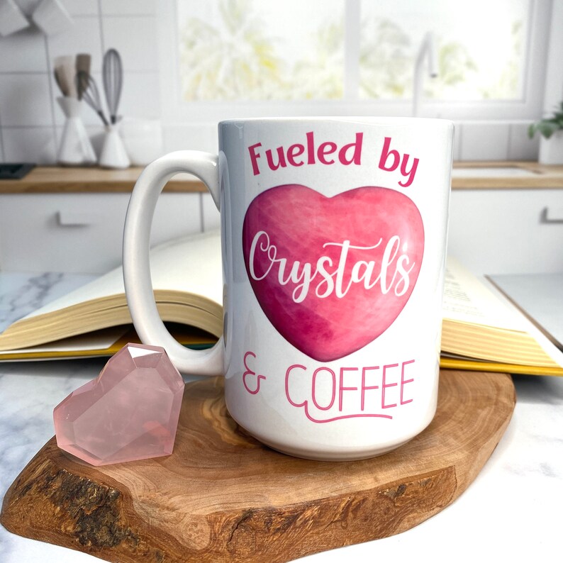 May include: White ceramic coffee mug with a pink heart and the text "Fueled by Crystals & Coffee".