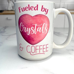 May include: White ceramic mug with a pink heart design and the text "Fueled by Crystals & COFFEE".