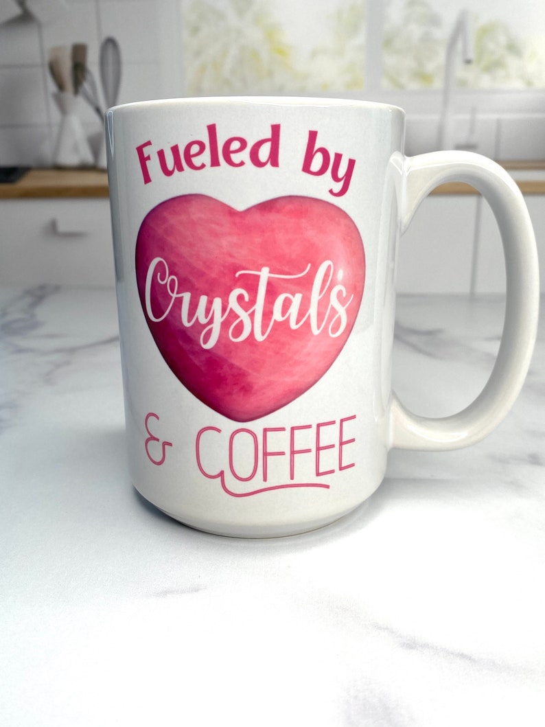 May include: White ceramic mug with a pink heart design and the text "Fueled by Crystals & COFFEE".