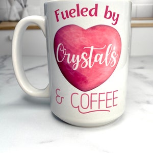 May include: White ceramic coffee mug with a pink heart and the text "Fueled by Crystals & Coffee".